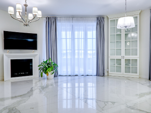 Bright modern living room with marble floors, a fireplace with a mounted TV, a potted plant, a glass-front cabinet, and sheer curtains over a large window.